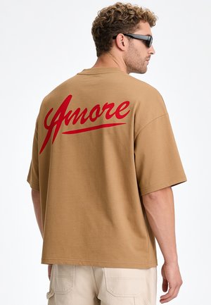 Uomo con capelli ricci e occhiali da sole che indossa una maglietta marrone con testo rosso "Amore" sulla schiena e pantaloni beige, in piedi di lato.