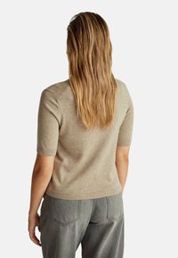 Frau mit langen nassen Haaren, die einen beigefarbenen kurzärmligen Pullover und graue Hosen trägt, steht mit dem Rücken zu einem schlichten Hintergrund.
