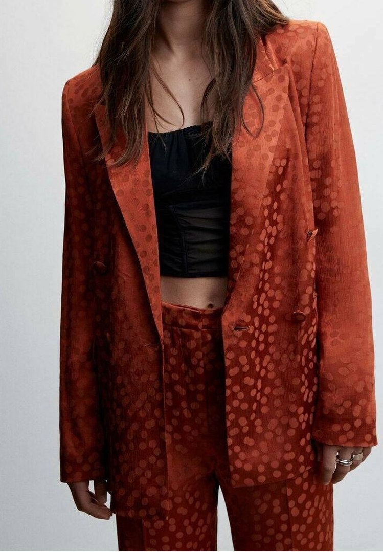 Femme portant un costume couleur rouille à motifs avec une veste croisée et un pantalon assorti par-dessus un crop top noir.