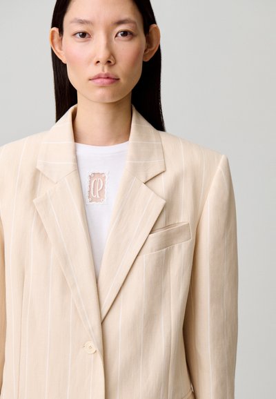 Blazer a righe beige con chiusura a un bottone, rever a incavo e taschino sul petto, indossato sopra una camicia bianca con dettaglio logo.