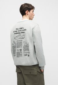 Νέος άνδρας που φοράει γκρι φούτερ με λεπτομερή μαύρα κείμενα και εικονογραφήσεις σχετικά με ένα αθλητικό παπούτσι στην πλάτη, συνδυασμένο με χακί cargo παντελόνια.