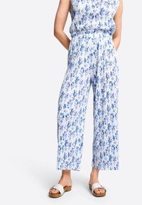 Witte stoffen jumpsuit met blauw bloemenpatroon, met een wijd uitlopende pijp, elastische tailleband, gecombineerd met witte sandalen en een lichtblauw top.