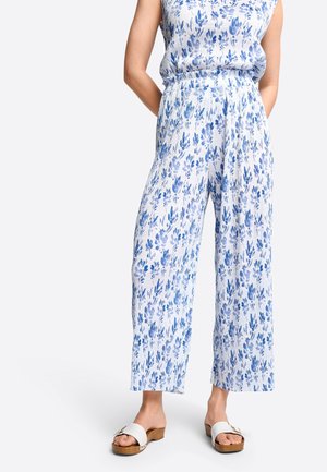 Witte stoffen jumpsuit met blauw bloemenpatroon, met een wijd uitlopende pijp, elastische tailleband, gecombineerd met witte sandalen en een lichtblauw top.