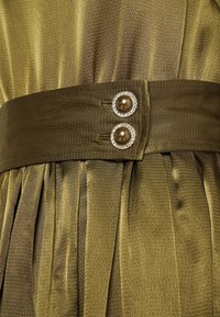 Tissu plissé vert olive avec une large ceinture assortie fixée par deux boutons ronds avec centres en perle et bordures en strass.