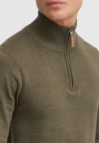 Pull en tricot vert olive avec un col haut côtelé et une fermeture éclair quart, présentant une étiquette en cuir près de la fermeture éclair. Texture lisse.