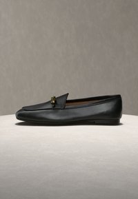 Μαύρο δερμάτινο μοκασίνι με μικρή χρυσή αγκράφα, επίπεδη σόλα και στρογγυλεμένη μύτη, τοποθετημένο σε ανοιχτή επιφάνεια με απλό φόντο.