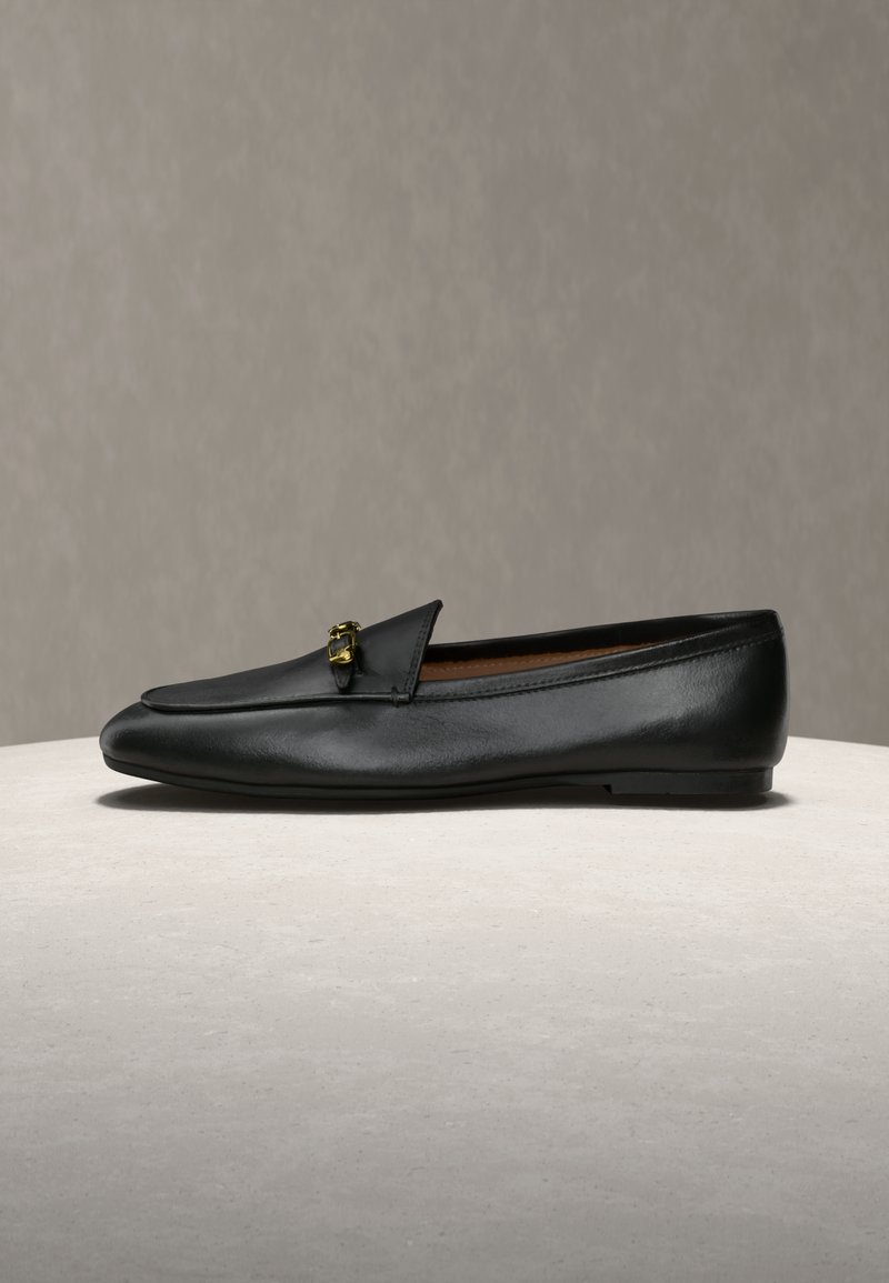 Μαύρο δερμάτινο μοκασίνι με μικρή χρυσή αγκράφα, επίπεδη σόλα και στρογγυλεμένη μύτη, τοποθετημένο σε ανοιχτή επιφάνεια με απλό φόντο.