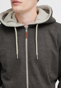 Sudadera gris con cremallera y capucha forrada de felpa, que incluye cordones de color crema, una cremallera plateada y una etiqueta de cuero marrón.