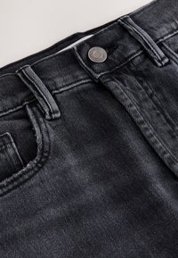 Nahaufnahme des Taillenbunds einer schwarzen Jeans aus Denim, die Knopf, Gürtelschlaufen und vordere Taschennaht zeigt.