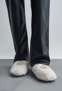 Fuzzy witte instapschoenen met rubberen zolen, voorzien van een cirkelvormig logoaccent. Gepaard met zwarte, glanzende leren broeken.