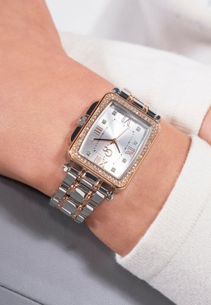 Ceas rectangular argintiu cu un bezel accentuat cu diamante, accente din aur roz și o curea din metal în două tonuri, purtat pe un încheietura cu o manșetă de culoare deschisă.