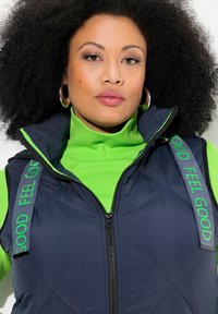 Granatowa pikowana kamizelka z zielonymi akcentami, z napisem "CZUJ SIĘ DOBRZE" na ramiączkach, noszona na jasnej zielonej golfie. Zamek błyskawiczny z przodu.