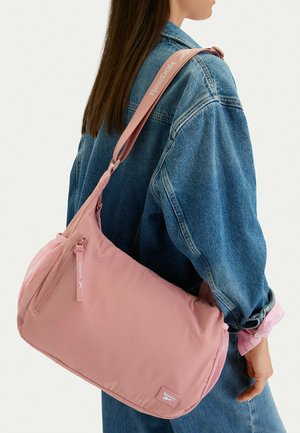 Mujer con chaqueta de mezclilla azul que lleva un bolso de hombro grande rosa de Reebok con bolsillo con cremallera y panel lateral de malla.