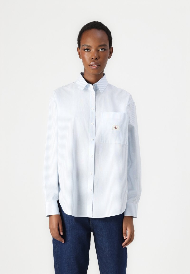 Striped blue and white long-sleeve shirt with a button-up collar, featuring a front pocket and embroidered logo detail on the pocket.