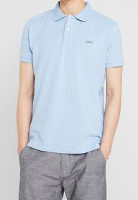 Ljusblå polo skjorta i bomull med ribbad krage och korta ärmar. Har en liten logotyp på bröstet. Kombineras med grå shorts.