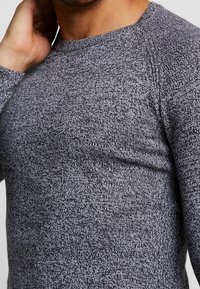 Grauer Strickpullover mit langen Ärmeln und strukturiertem, gesprenkeltem Muster. Verfügt über einen Rundhalsausschnitt und Raglanärmel. Weicher Stoff, figurbetonter Schnitt.