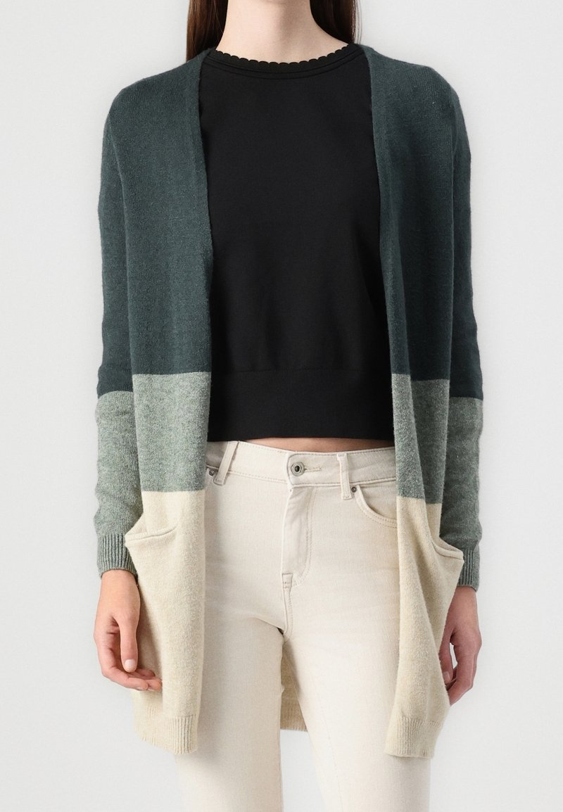 Femme portant un long cardigan trois tons en vert foncé, gris et beige sur un haut noir et un pantalon beige clair, mains dans les poches du cardigan.
