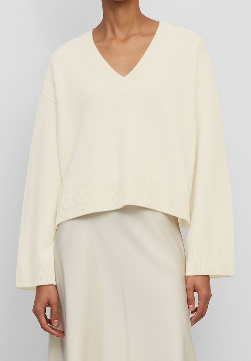 Pull à col en V couleur crème avec poignets et ourlet côtelés, présentant une coupe ample et courte avec une texture douce. Porté sur une jupe assortie crème.