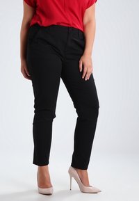 Pantaloni neri aderenti con una texture liscia, dotati di tasche laterali e chiusura a bottone, abbinati a tacchi alti rosa chiaro e una blusa rossa.