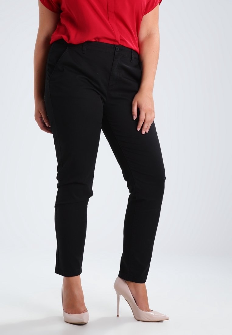 Pantaloni neri aderenti con una texture liscia, dotati di tasche laterali e chiusura a bottone, abbinati a tacchi alti rosa chiaro e una blusa rossa.