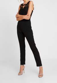 Mono negro con detalles de encaje en el escote, piernas ajustadas y tirantes ajustables. Combinado con tacones altos plateados con tiras.