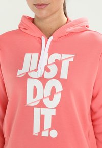Růžová mikina s kapucí vyrobená z měkké tkaniny. Má bílou grafiku s textem "JUST DO IT" ve zvýrazněném, rozděleném designu. Disponuje šňůrkou na stažení kapuce.