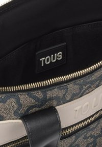 Bolso de lona con un patrón floral azul, detalles en cuero negro y cierre de cremallera plateado. Logo "TOUS" grabado en el interior.