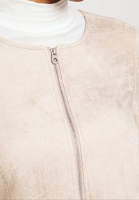Gros plan d'une veste en daim beige avec une fermeture éclair argentée portée sur un haut blanc à col montant.