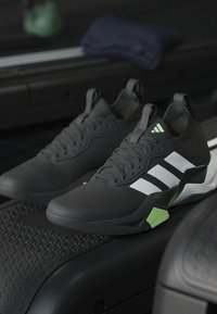 Paire de chaussures de sport Adidas noires avec des bandes blanches et des accents verts placée sur une surface de tapis roulant dans une salle de sport.
