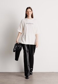 Υπερμεγέθυ λευκό t-shirt με το κείμενο "Calvin Klein", συνδυασμένο με λαμπερές, μαύρες πλισέ παντελόνες και ένα μαύρο μπουφάν ριγμένο στον έναν βραχίονά.