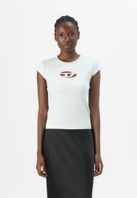 Tricou ajustat, cu maneci scurte, de culoare albastru deschis, cu un logo oval roz pe piept, purtat cu o fustă neagră. Material neted și design minim.