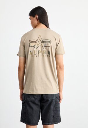 Beige Baumwoll-T-Shirt mit einem grafischen Design, das die Worte "ALPHA INDUSTRIES" in dunklen Farben und ein emblemartiges Motiv auf der Rückseite zeigt.