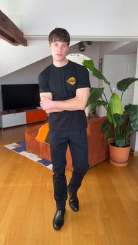 Mladý muž v čiernom tričku a nohaviciach stojaci so založenými rukami v modernom obývacom priestore s veľkou rastlinou a koralovou pohovkou.