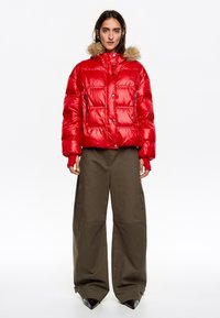 Czerwona kurtka puchowa z kapturem obszytym futrem, błyszcząca tkanina, boczne zamki błyskawiczne i krótki fason, zestawiona z luźnymi oliwkowozielonymi spodniami.