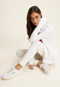 Chaqueta blanca con cierre y acentos dorados, camiseta burdeos, pantalones jogger crema y zapatillas blancas, dispuestos en una pose sentada contra un fondo claro.