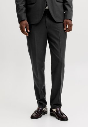 Férfi fekete szabott nadrágot, fekete blézert, fehér zoknit és sötétbarna bojtos mokaszint visel, fehér háttér előtt áll.