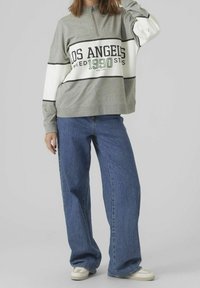 Grijze half-zip sweatshirt met een wit en zwart streep, voorzien van de tekst "LOS ANGELES 1990"; gecombineerd met losse blauwe denim jeans en witte sneakers.
