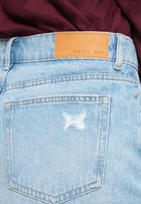 Ανοιχτό μπλε τζιν παντελόνι με καφέ δερμάτινη ετικέτα στο πίσω μέρος που γράφει «NOISY MAY». Διαθέτει μικρό, ξεφτισμένο σχέδιο σταυρού στην τσέπη.