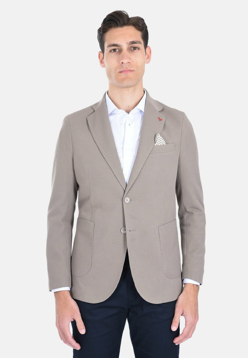Blazer beige testurizzato con due tasche frontali, due bottoni e un fazzoletto bianco. Indossato sopra una camicia bianca con un accento rosso.