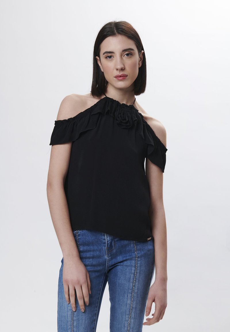 Top negro sin mangas con diseño de volantes y acento floral en el escote, combinado con jeans de denim azul de corte ajustado.