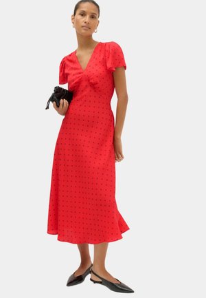 Kvinde i en rød midi-kjole med små sorte prikker, pufærmer, der holder en lille sort håndtaske, iført sorte spidse slingback-flats.