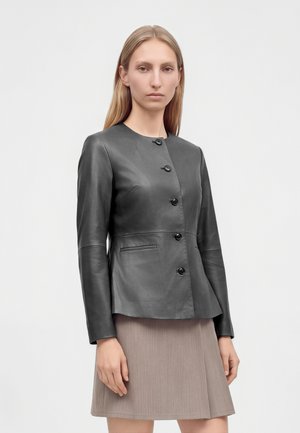 Femme portant une veste en cuir noir ajustée avec des boutons et une jupe beige longueur genou, debout devant un fond blanc uni.