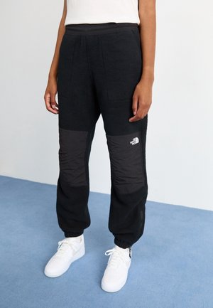 Person iført sorte fleecebukser med forstærkede knælapper og hvide sneakers, stående på et blåt tæppebelagt gulv.