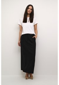 Jupe longue noire avec des poches latérales, associée à un t-shirt blanc à col rond. Le mannequin porte des cheveux noirs raides et des escarpins beiges à bout fermé.