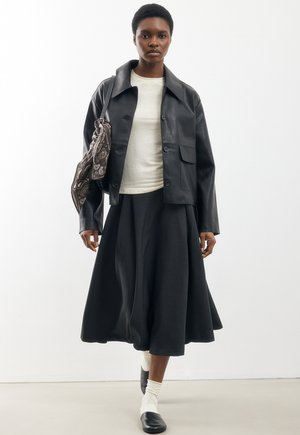 Kobieta ubrana w czarną skórzaną kurtkę, biały top, czarną spódnicę midi, białe skarpetki i czarne buty, trzymająca brązową torebkę z wzorem na białym tle.