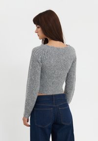 Szary, dziany krótki sweter z długimi rękawami, o prążkowanej fakturze. Noszony z ciemnoniebieskimi spodniami na wysokim stanie, widok z tyłu.