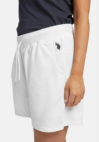 Białe bawełniane szorty z prążkowanym pasem, sznurkiem do wiązania, bocznymi kieszeniami i małym czarnym haftowanym logo po lewej stronie.