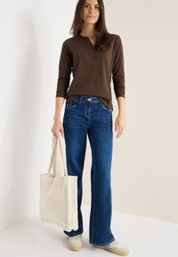 Braunes langärmliges Henley-Shirt, blaue Schlagjeans und eine weiße strukturierte Tote-Tasche. Das Outfit wird von hellen Sneakers mit beiger Sohle ergänzt.