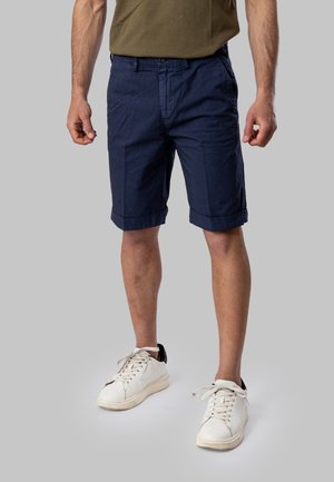 Uomo che indossa pantaloncini blu navy fino al ginocchio, scarpe da ginnastica bianche e una camicia verde oliva, in piedi davanti a uno sfondo grigio.