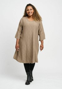 Beige långärmad klänning med en texturerad mönster, rund halsringning och en löst sittande silhuett, kombinerad med svarta leggings och ankelstövlar.